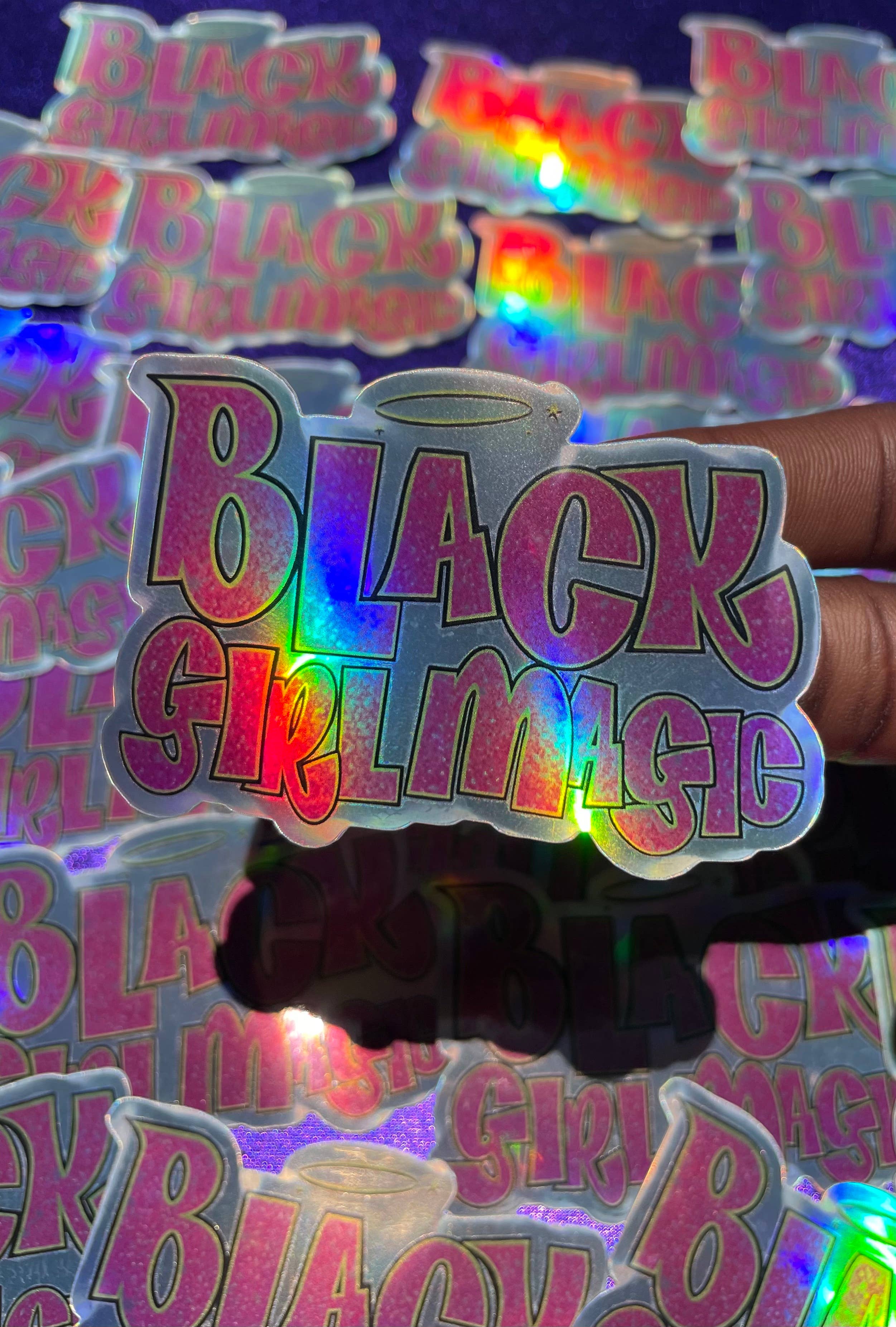 A vinyl sticker with the phrase 'Black Girl Magic' written in pink and purple colors, held against a blurred background of similar stickers.